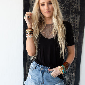 A woman with long blonde hair wears the Scoop Neck Bralette Tee in black, paired with ripped denim shorts and multiple bracelets. She stands before a decorative dark tapestry, showcasing effortless boho style.
