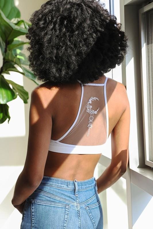 A woman with curly hair stands indoors by a window, sunlight streaming in as she faces away. She wears the Crescent Moon Tattoo Bralette - White, featuring crescent moon details on the sheer back panel, paired with high-waisted blue jeans.
