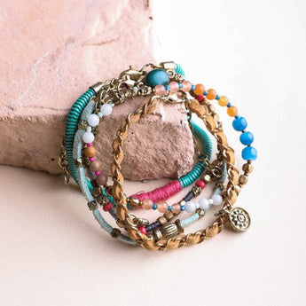 The Sunburst Stacked Bracelet Set - Multi features a lively mix of colorful beaded and braided boho bracelets with charms in blue, pink, turquoise, white, and gold, displayed on a light stone surface.