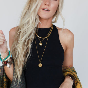 A woman with long, wavy blonde hair wears a black High Neck Racerback Brami, layered gold necklaces, several bracelets, and a patterned shawl over one arm as she stands against a plain light background.