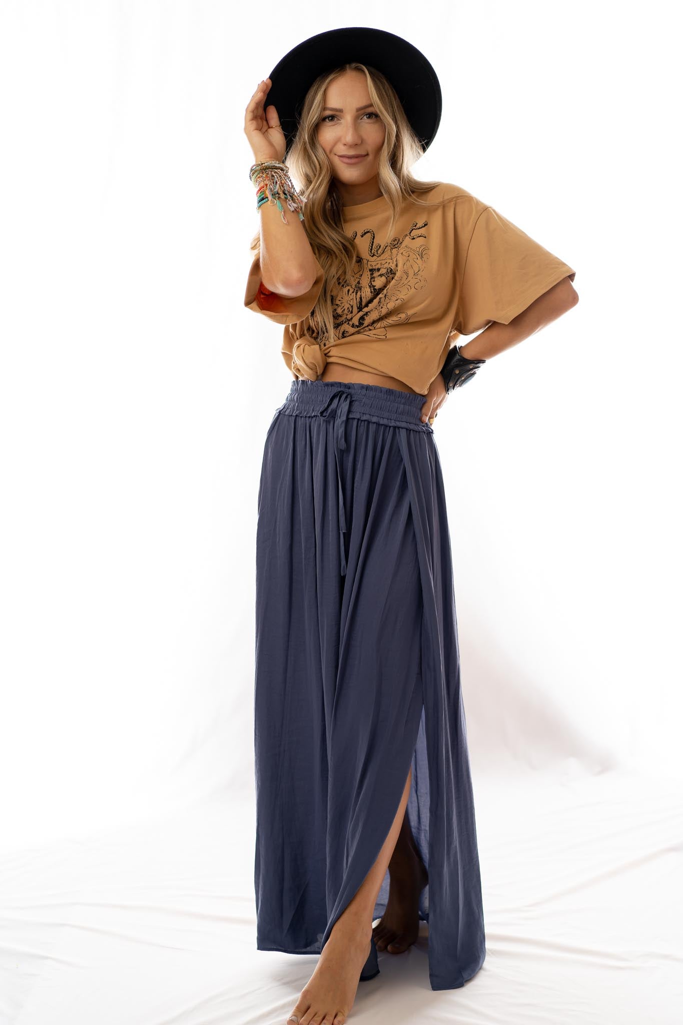 Woman poses barefoot against a white background, wearing a wide-brimmed black hat, tan graphic tee knotted at the waist, and the Stasia Maxi Skirt - Blue with side slits, styled with colorful bracelets.