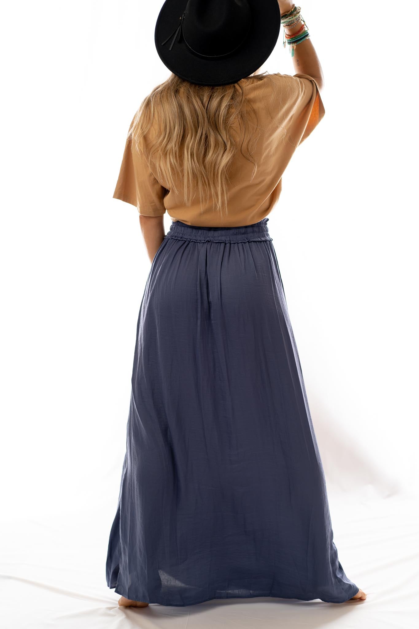 A woman with long wavy blonde hair, barefoot on a white background, wears a tan top, multiple bracelets, a black wide-brim hat, and the Stasia Maxi Skirt – Blue, styled high waisted with her back to the camera.