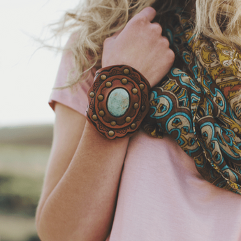 A woman with long curly blonde hair and a light pink t-shirt wears a colorful scarf, showing off the Aegean Boho Leather Cuff Bracelet - Brown on her wrist.