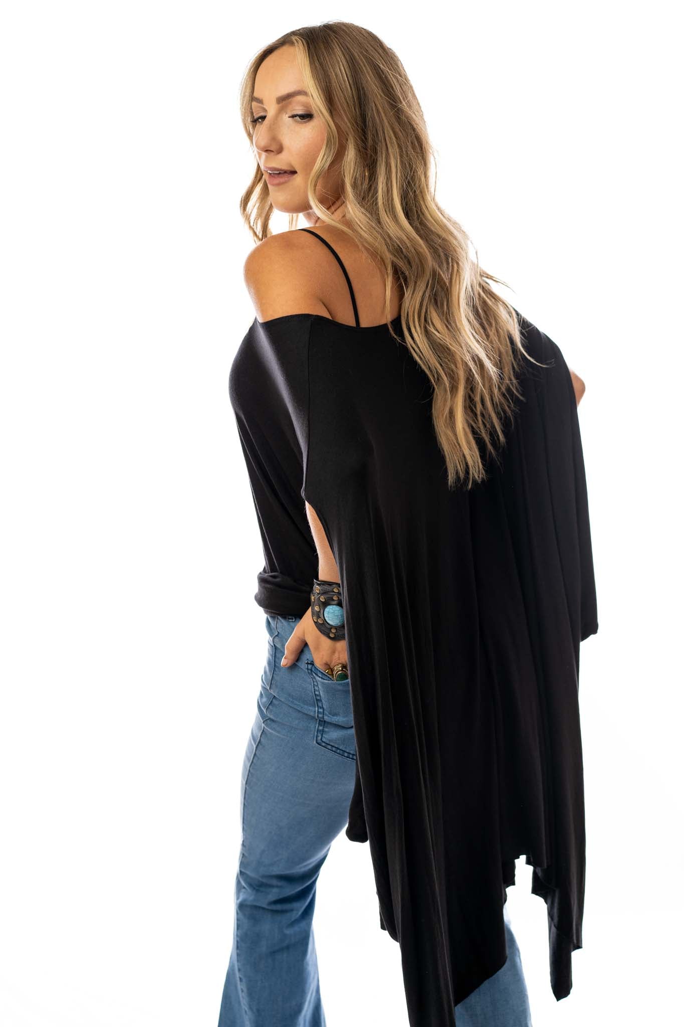 A woman with long, wavy blonde hair wears The Wren Tunic - Black styled as a relaxed off-the-shoulder top with blue jeans, standing sideways and looking back against a white background.