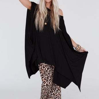 A woman with long blonde hair poses against a plain background wearing The Wren Tunic - Black, leopard print flared pants, a knitted beige hat, and multiple bracelets for a relaxed, stylish look.