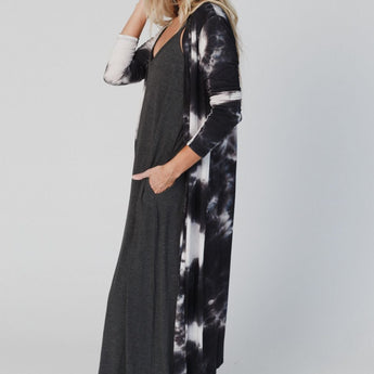A woman with long blonde hair wears the My Go To Duster Cardigan - Black Tie Dye over a black maxi dress. She stands sideways, looking at the camera with one hand in her pocket and the other touching her hair.