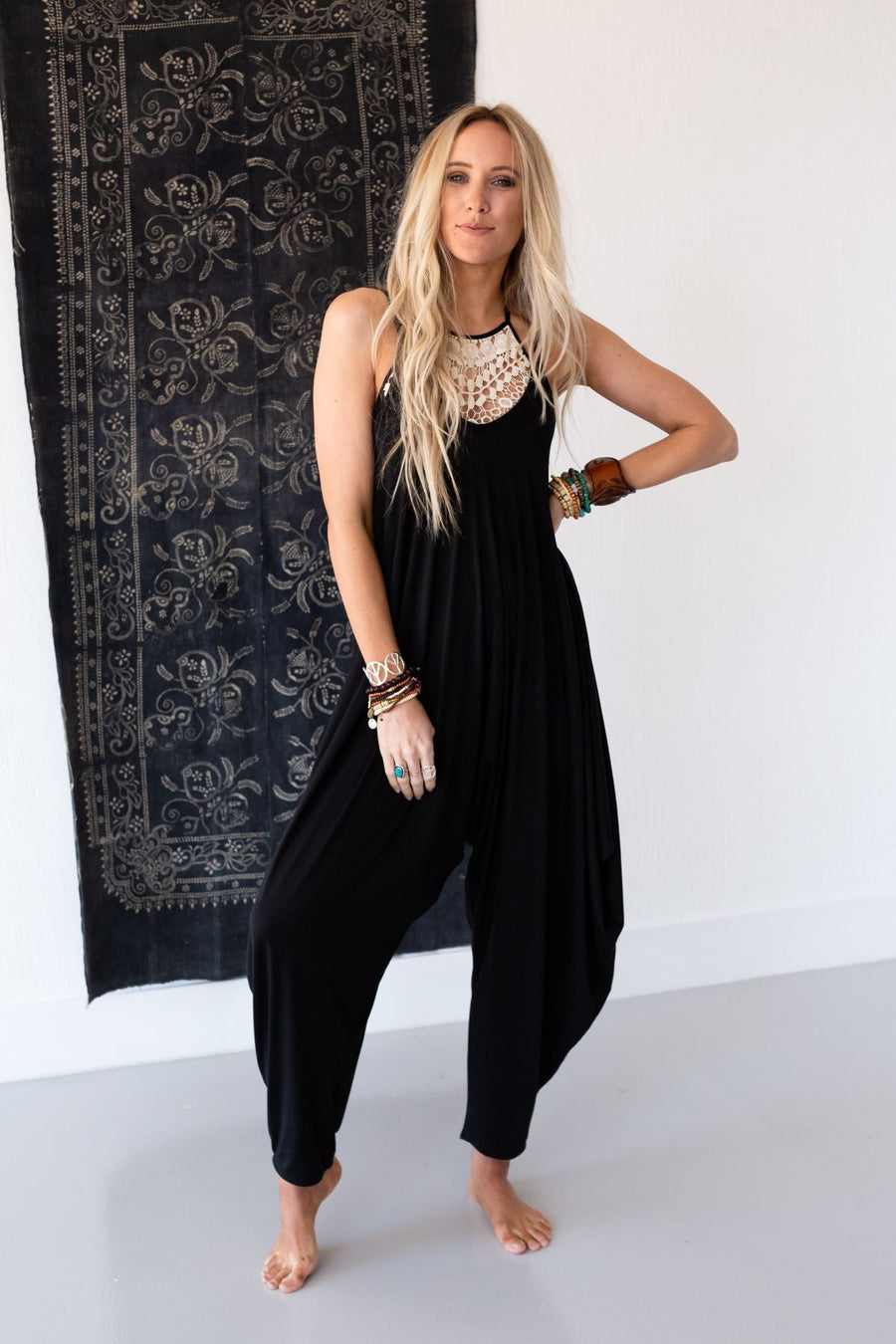 A woman with long blonde hair stands barefoot indoors, confidently posing in The Perfect Harem Jumpsuit - Black with a white crochet neckline. She accessorizes with bracelets and rings in front of a decorative black tapestry.