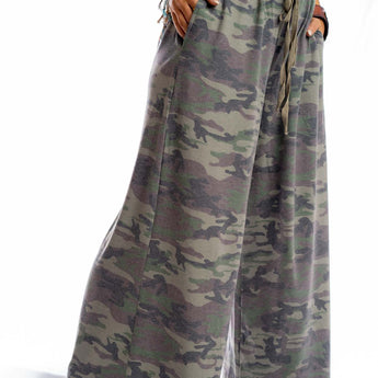 A person wears the Calling All Camo Lounge Pants - Olive with a white knotted top, standing barefoot on a white surface, hands in pockets and colorful bracelets on their wrist.
