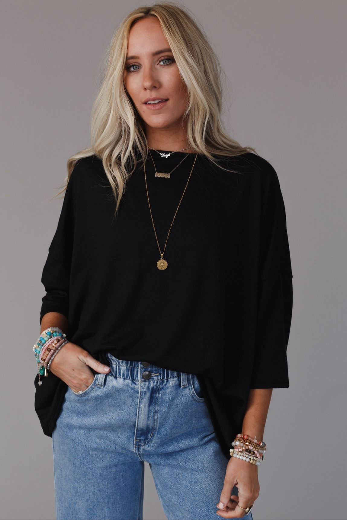 A woman with long blonde hair wears The Nest Cozy Cool Oversized Tee in black, paired with light blue jeans, layered necklaces, and colorful beaded bracelets, standing with one hand in her pocket against a neutral background.