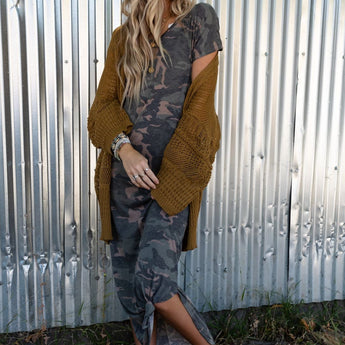 A woman with long blonde hair stands by a corrugated metal wall, wearing the Call It Comfort Tee Dress - Camo with a knotted hem, a chunky brown cardigan, stacked bracelets, and studded sandals as she looks to the side.