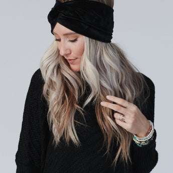 A woman with long, wavy blonde hair wears the Everyday Velvet Headband - Black with criss-cross detailing and a black sweater. She looks down, touching her hair and displaying several bracelets and rings against a light background.
