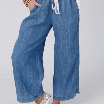 Wearing the So Comfy Wide Leg Denim Pant - Denim with an elastic drawstring waist, a white top, checkered slip-on shoes, and colorful bracelets, a person stands confidently with one hand in their pocket.