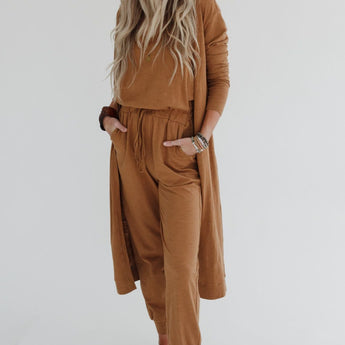 A woman with long, wavy blonde hair stands barefoot against a white background, wearing the Set to Go Textured Jumpsuit And Cardigan Set in camel, with her hands in her pockets as she looks off to the side.