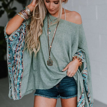 A woman wearing the Fallon Embroidered Sleeve Poncho in sage, featuring colorful sleeves, styled with layered necklaces, bracelets, and denim shorts, stands indoors looking down and touching her hair.