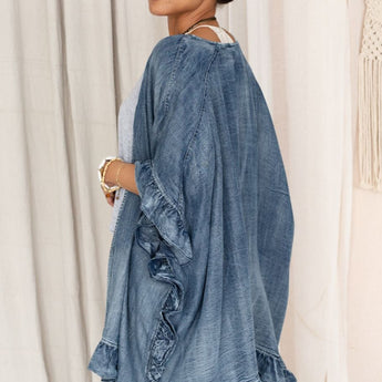 A woman with dark hair pulled back wears the Robbie Denim Jacket - Blue, an oversized fit with ruffle trim, over a grey top and jeans. She stands sideways looking over her shoulder, with neutral curtains in the background.