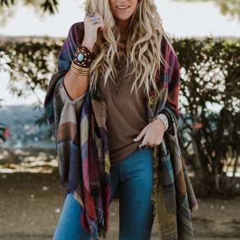 A woman with long, wavy blonde hair smiles outdoors in a Cozy Plaid Pocket Ruana Wrap - Fuchsia over her brown top and blue jeans, accessorizing with bracelets and rings amid lush bushes and trees.