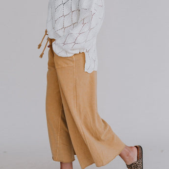 A person in a white patterned sweater, Relaxing Robin Wide Leg Pant - Mustard, and leopard print slide sandals walks against a plain light grey background. Their face is not visible.