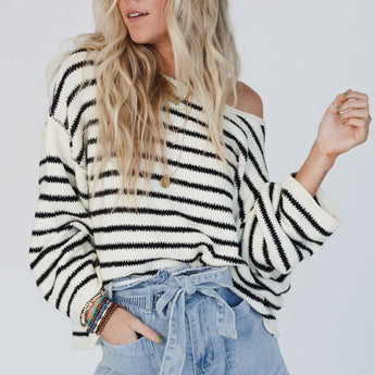 A woman with long, wavy blonde hair wears the All My Stripes Oversized Sweater in cream and black off one shoulder, paired with high-waisted, tie-front denim shorts, standing against a plain light background.