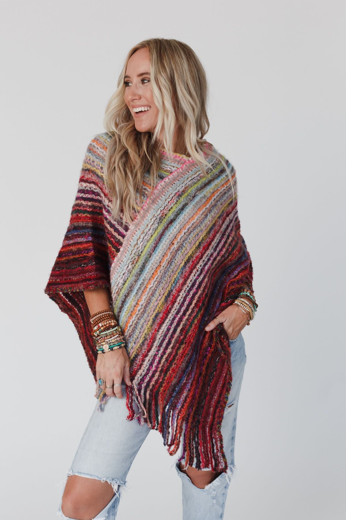 A woman with long blonde hair smiles to the side, modeling the Madeleine Poncho - Multi. She wears ripped light-wash jeans, several bracelets, and stands against a plain white background.