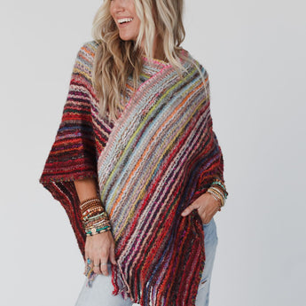 A woman with long blonde hair smiles to the side, modeling the Madeleine Poncho - Multi. She wears ripped light-wash jeans, several bracelets, and stands against a plain white background.