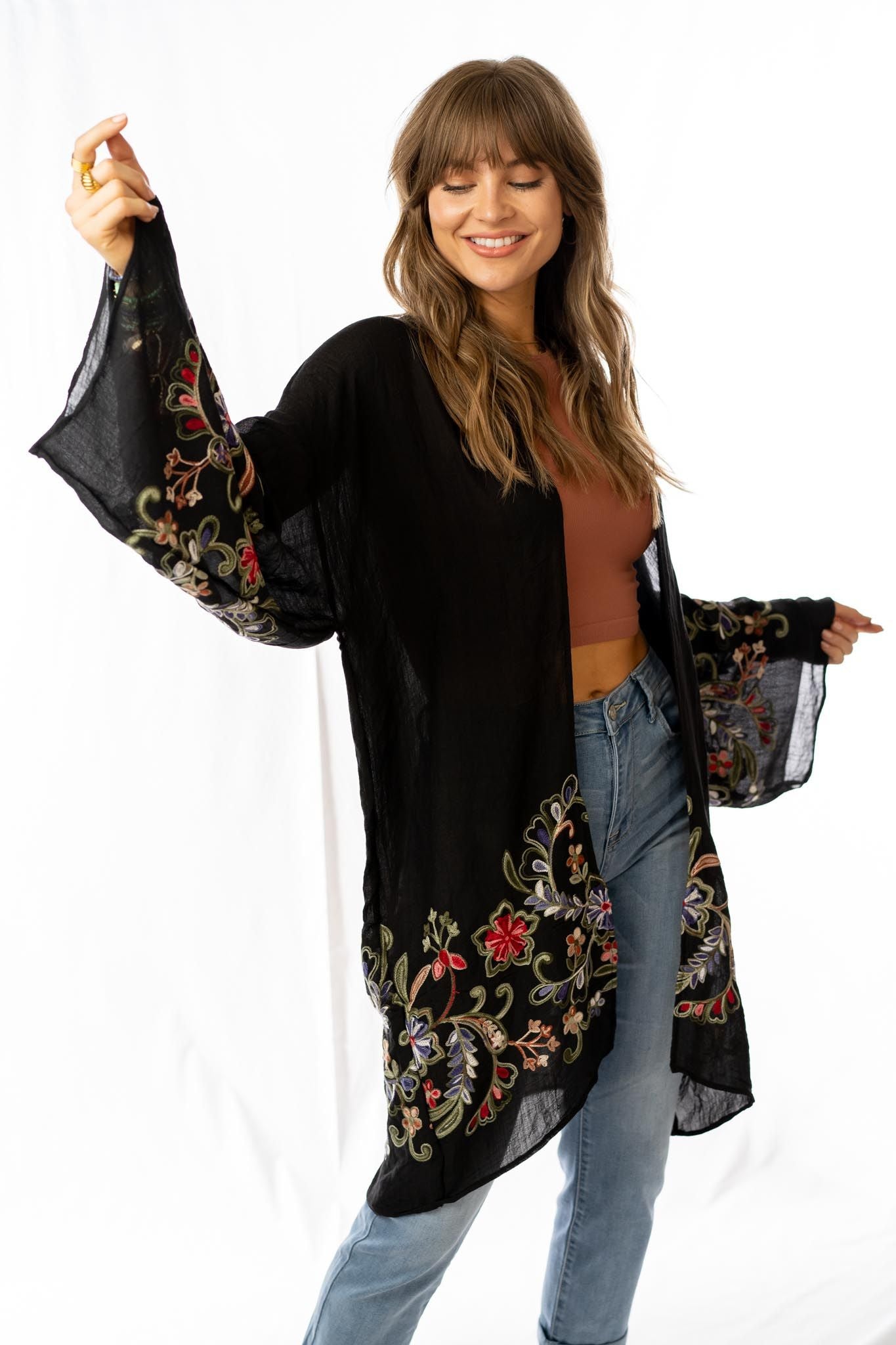 A smiling woman in the Fun In Florals Kimono - Black, featuring colorful embroidery, brown crop top, and blue jeans poses with one arm raised against a white background.