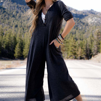 A smiling woman stands on a sunny mountain road in the Simply Comfort Jumpsuit - Dark Gray, a scoop neck boho style with an oversize silhouette. She pairs it with white sandals and colorful bracelets, framed by pine trees and rolling hills.