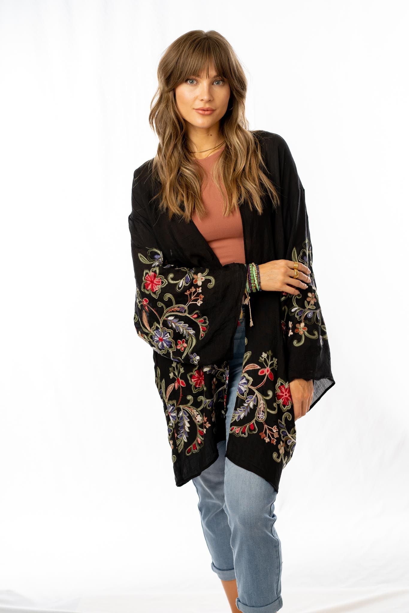 A woman with long, wavy brown hair stands against a white background, wearing cuffed jeans, a rust-colored top, and the Fun In Florals Kimono - Black with colorful embroidery. She poses with one hand on her hip and looks relaxed.