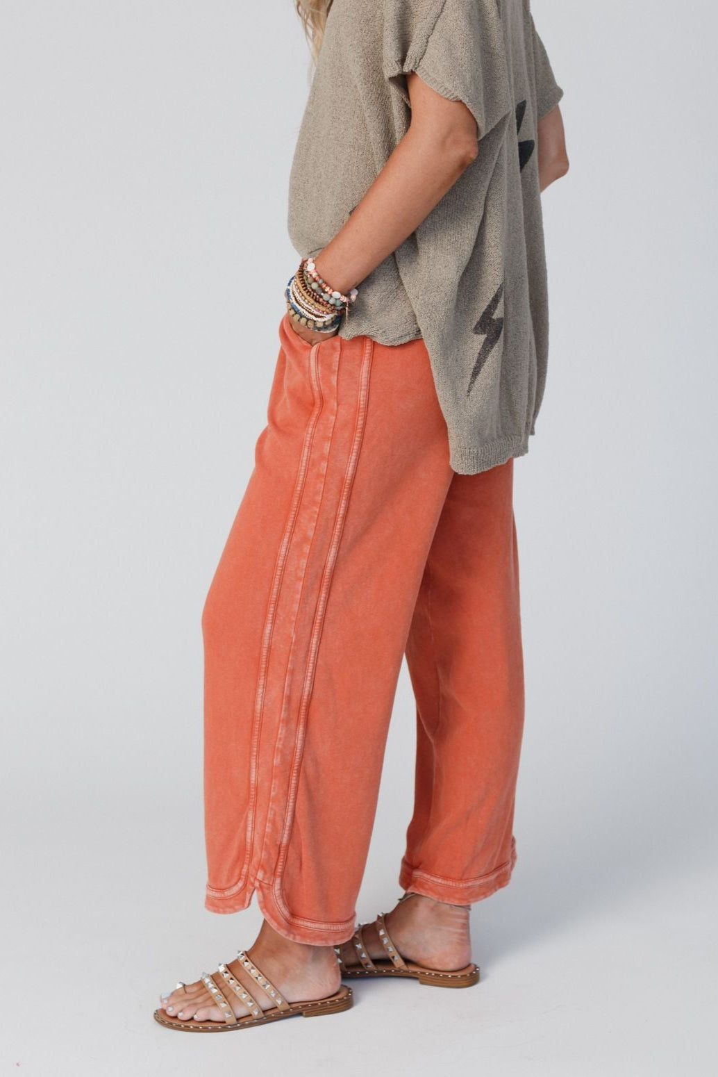 A person stands sideways wearing So Comfy Wide Leg Pant Cropped Length - Faded Rust with a high rise elastic waist, a loose taupe lightning bolt shirt, beaded bracelets, and studded sandals. Their face is not visible.