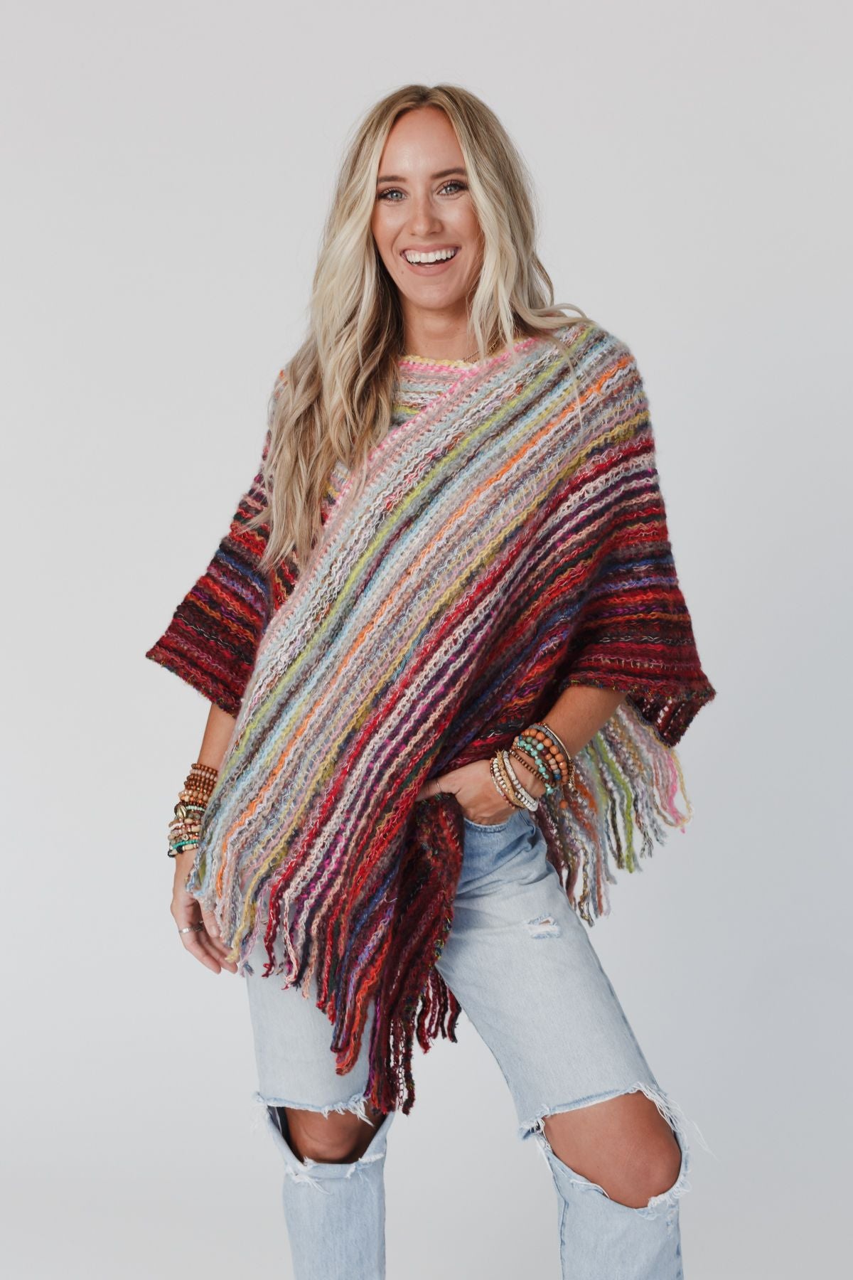 A woman with long blonde hair smiles while wearing the Madeleine Poncho - Multi, a colorful striped knit with boho fringes, paired with ripped light blue jeans and several bracelets. She stands against a plain white background.