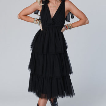 A woman with shoulder-length blonde hair stands with hands on hips, wearing the sleeveless Boho Ballerina Tiered Tulle Dress in black, styled with bow straps, colorful bracelets, and black cowboy boots against a neutral background.