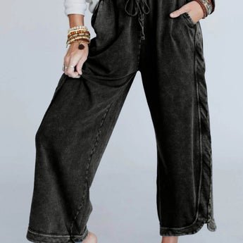 A person stands barefoot, hands in pockets, wearing a white long-sleeve top and the So Comfy Wide Leg Pant Cropped Length - Charcoal with a high-rise elastic waist, accessorized with multiple bracelets on both wrists.