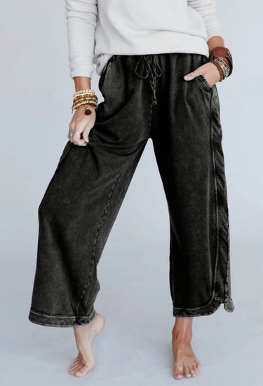 A person stands barefoot, hands in pockets, wearing a white long-sleeve top and the So Comfy Wide Leg Pant Cropped Length - Charcoal with a high-rise elastic waist, accessorized with multiple bracelets on both wrists.