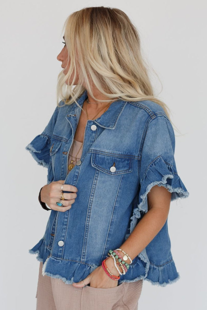 A woman with long blonde hair wears the Scottie Ruffle Denim Jacket - Light Blue, featuring ruffled sleeves and a cropped hemline. She pairs it with light brown pants and colorful bracelets, standing sideways against a plain white background.