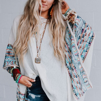 A woman with long, wavy blonde hair wears the Fallon Embroidered Sleeve Poncho in ivory, paired with denim shorts, layered necklaces, and multiple bracelets, standing against a light gray brick wall.