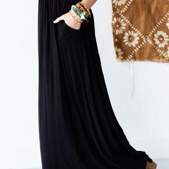 A person wearing The Perfect Pocketed Maxi Skirt - Black, a sleeveless white top, beaded bracelets, and brown ankle boots stands next to a brown patterned textile hanging on a white wall.