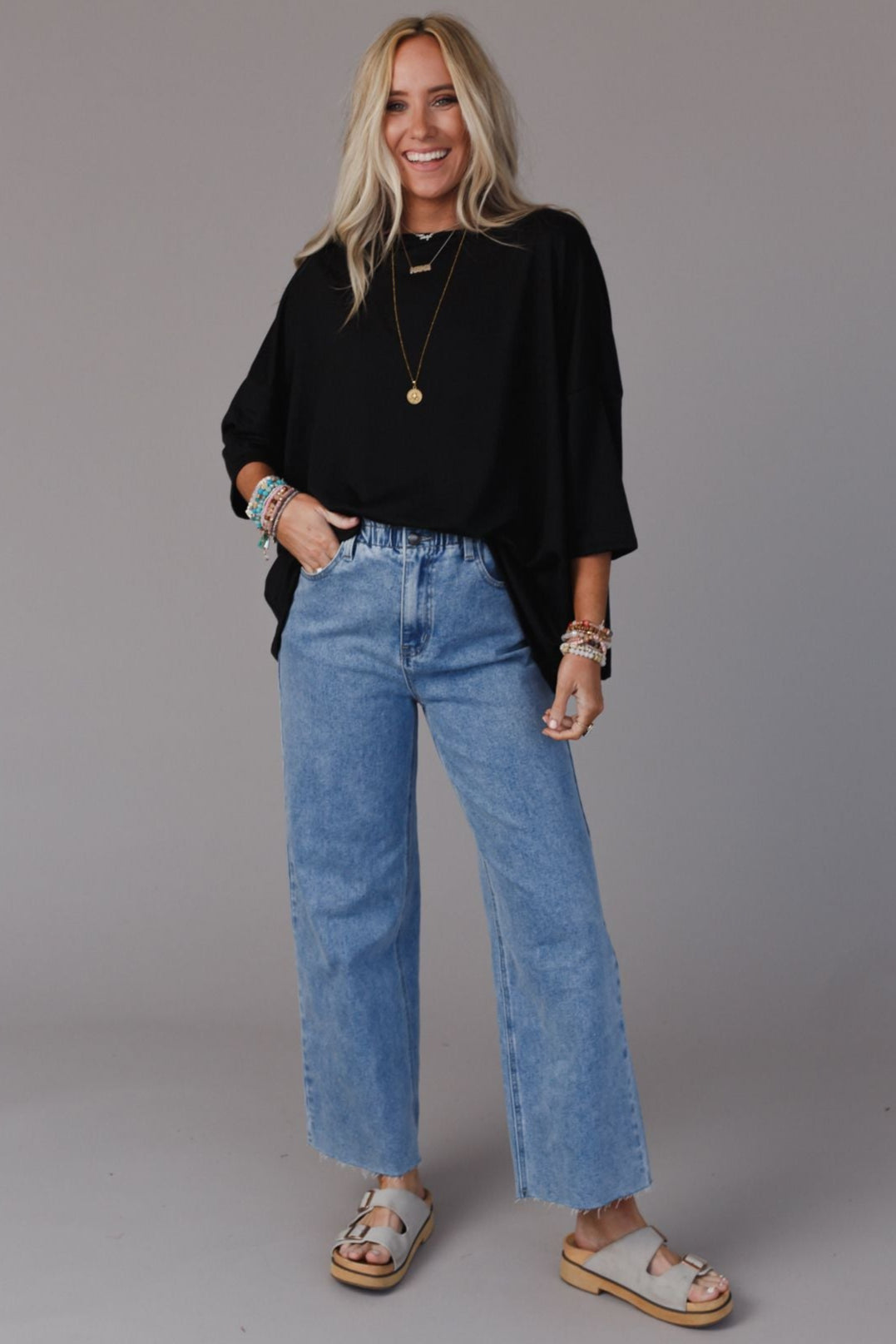 A smiling woman with long blonde hair wears The Nest Cozy Cool Oversized Tee in black, paired with light blue high-waisted wide-leg jeans, beige sandals, and layered necklaces against a plain gray background.