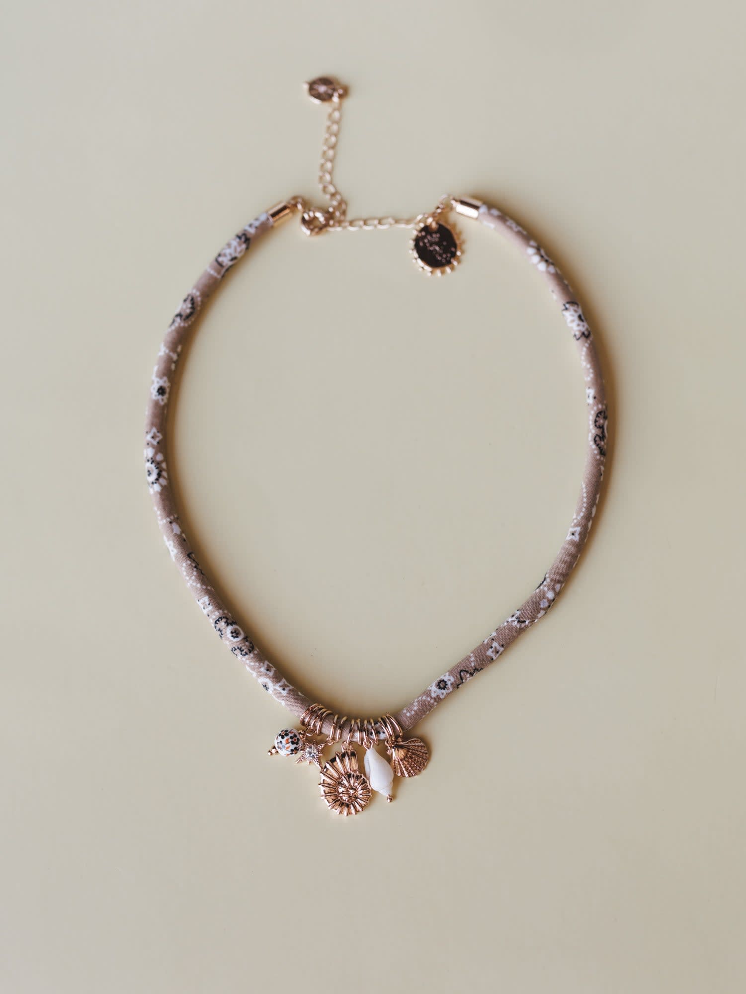 A fabric-wrapped choker necklace in dusty mauve with a delicate floral print, layered with rose gold ocean-inspired charms — shells, a sunburst, and a white stone drop — laid flat on a warm sage background.