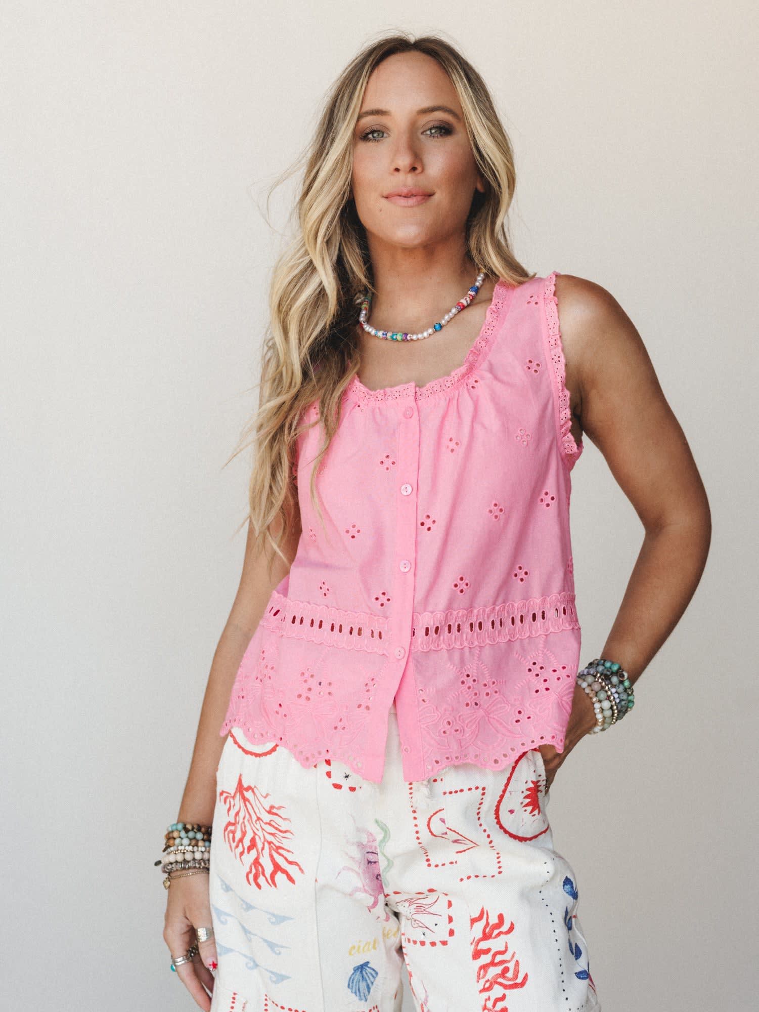 A bright pink eyelet button-front tank top styled with illustrated white pants and layered beaded jewelry — playful and summery with a handcrafted, free-spirited feel.