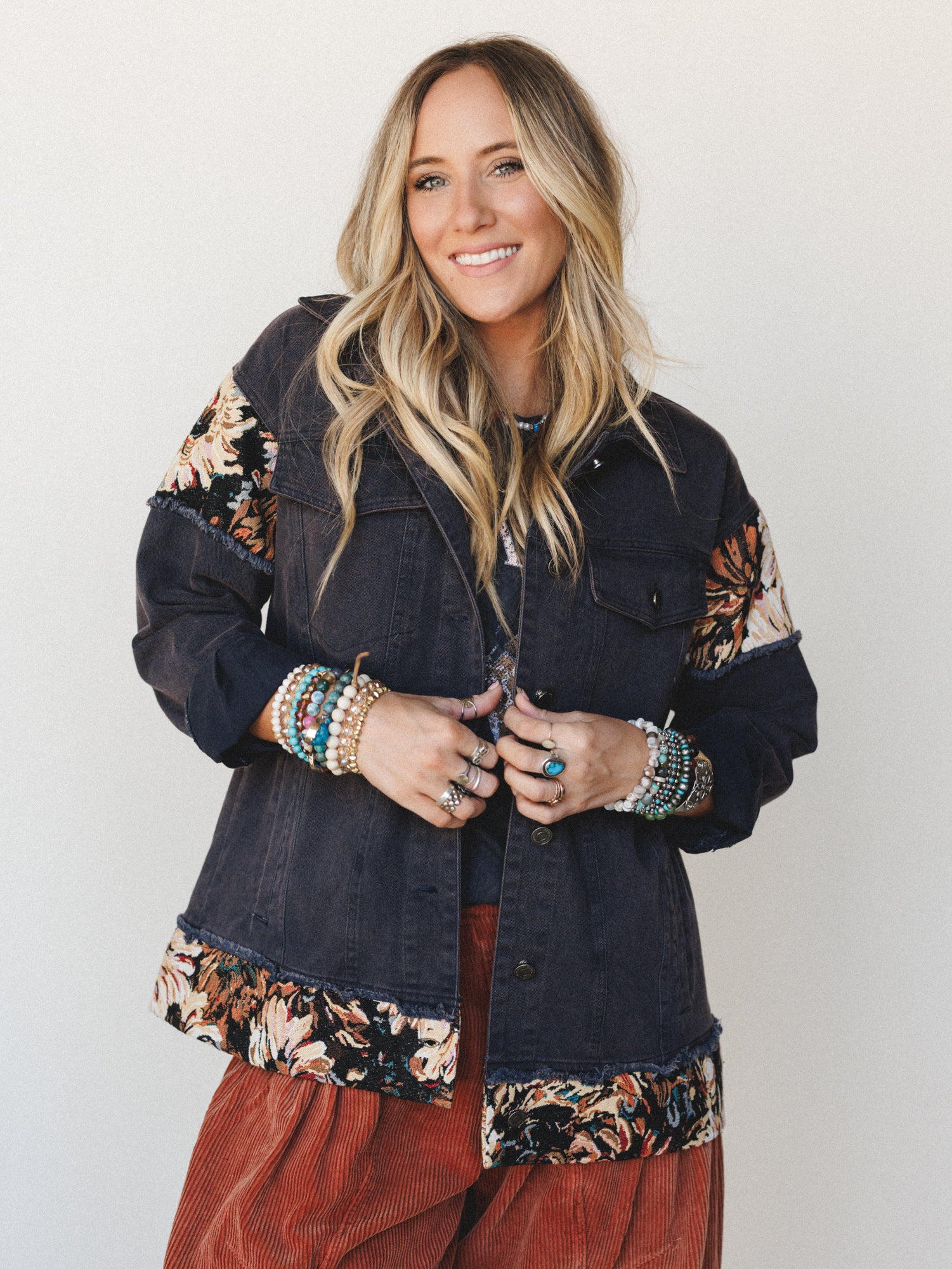A smiling model layers a washed navy denim jacket with tapestry floral patchwork panels and frayed trim over a rust corduroy skirt, styled with stacked beaded bracelets and silver rings for a lived-in boho feel.