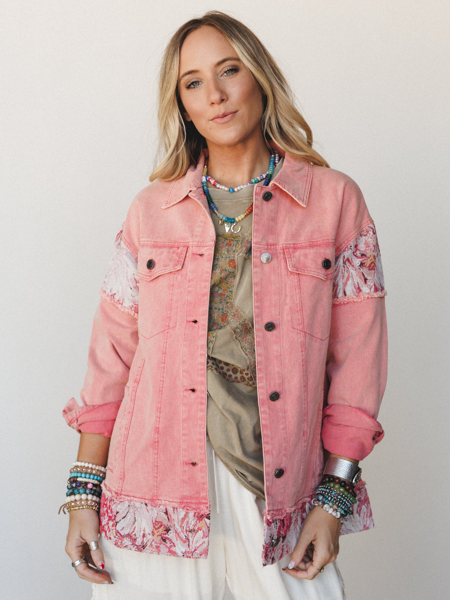 A pink acid-wash denim jacket with floral patchwork panels at the shoulders and hem, layered over a graphic tee and white pants — the kind of piece that makes a whole outfit feel intentional without trying too hard.