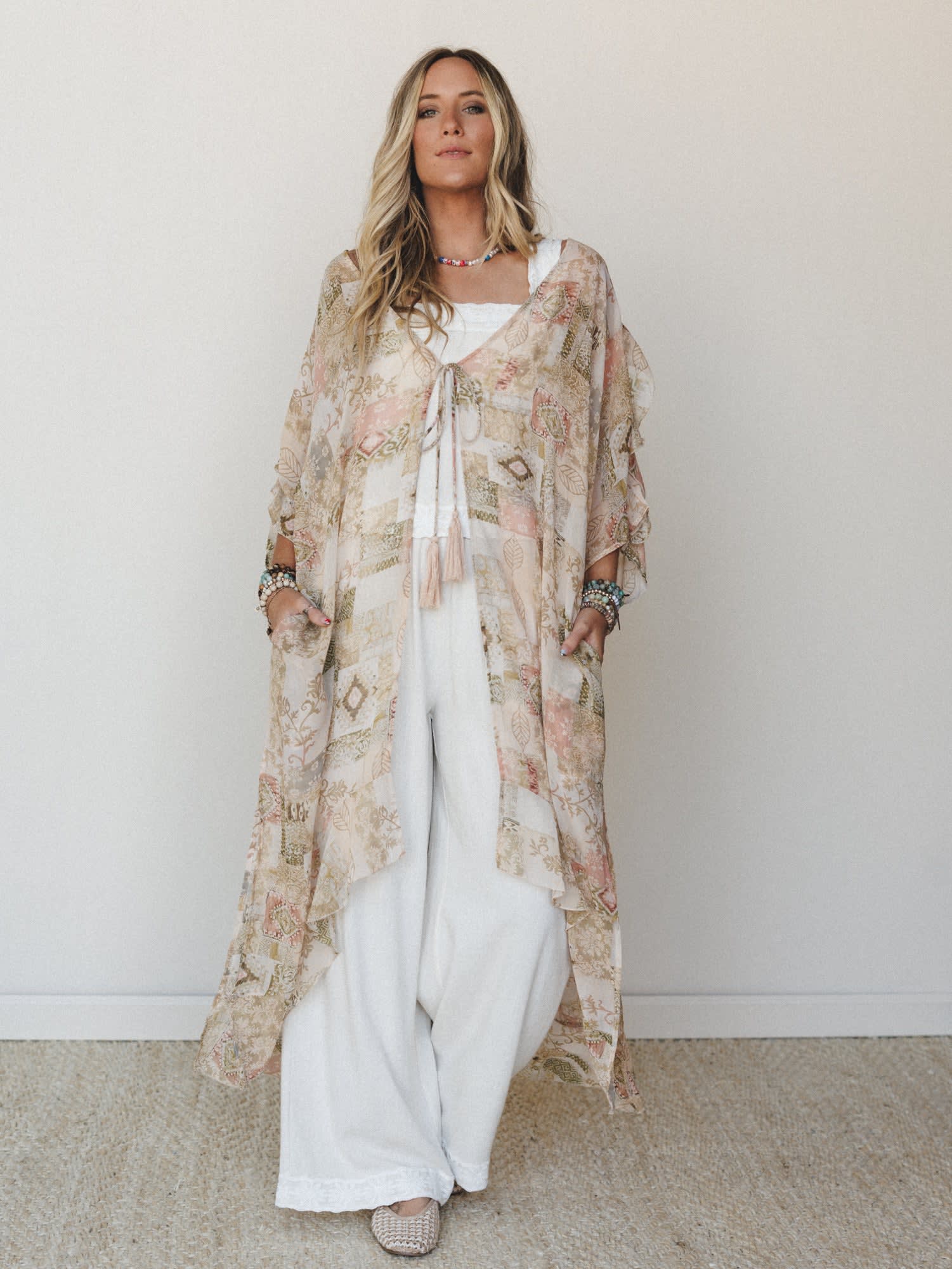 A flowy maxi kimono in a soft patchwork-inspired botanical print layers effortlessly over wide-leg white pants and a white cami, styled with stacked bracelets and a beaded necklace for a look that feels like a really good afternoon.