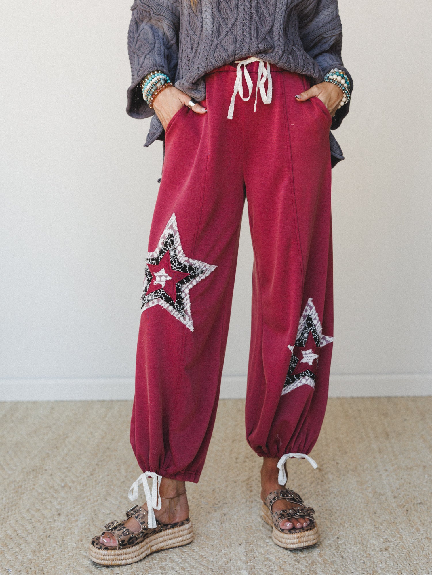 A relaxed wide-leg jogger in deep magenta with layered star patchwork appliqués on each leg, styled with a chunky cable-knit sweater, stacked beaded bracelets, and leopard print platform sandals — the kind of outfit that's effortlessly expressive wit