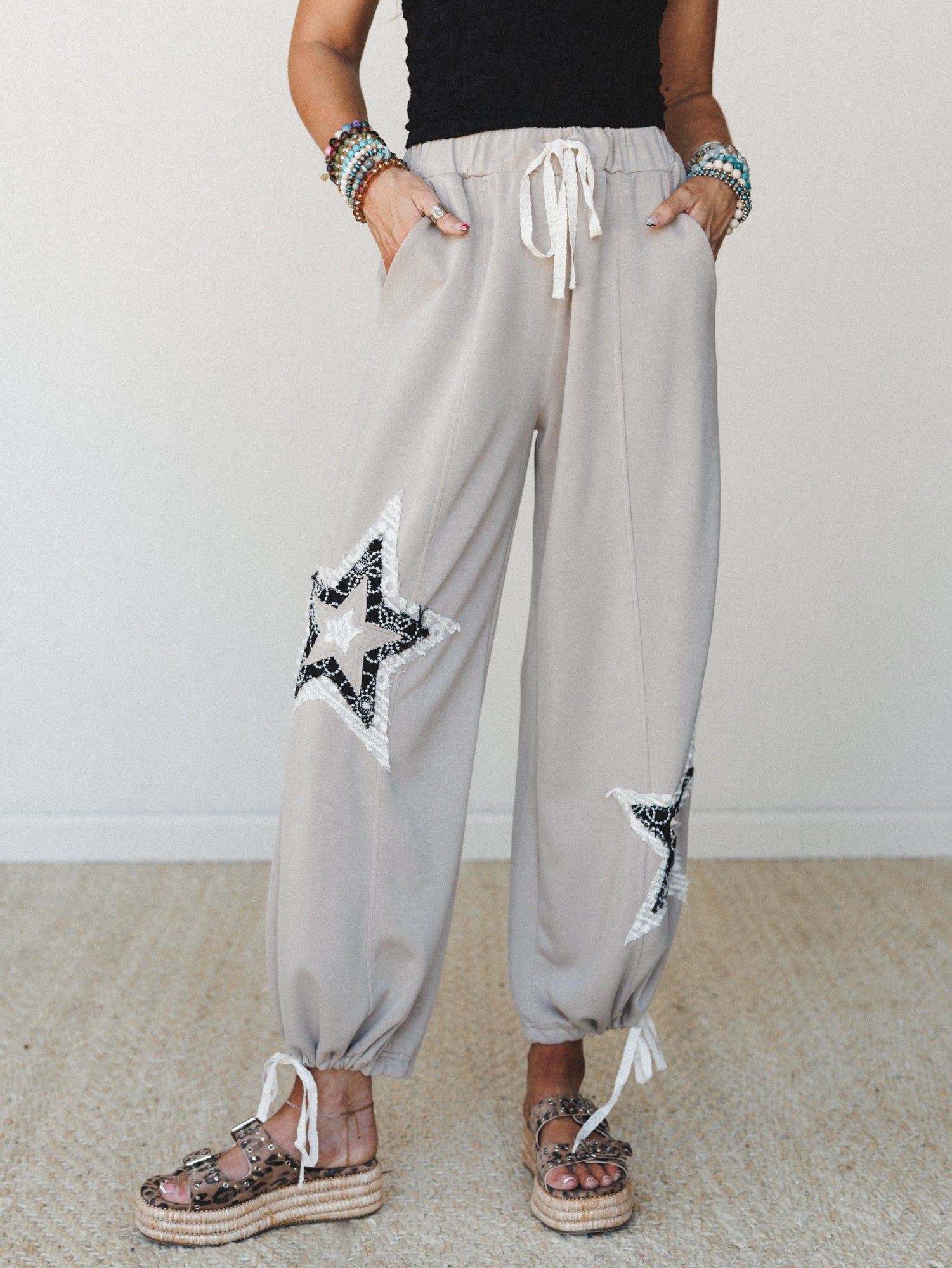 Taupe jogger-style pants with lace-edged star appliqués on each thigh, styled with a black textured top, stacked beaded bracelets, and leopard print platform sandals — casual and expressive with a boho edge.