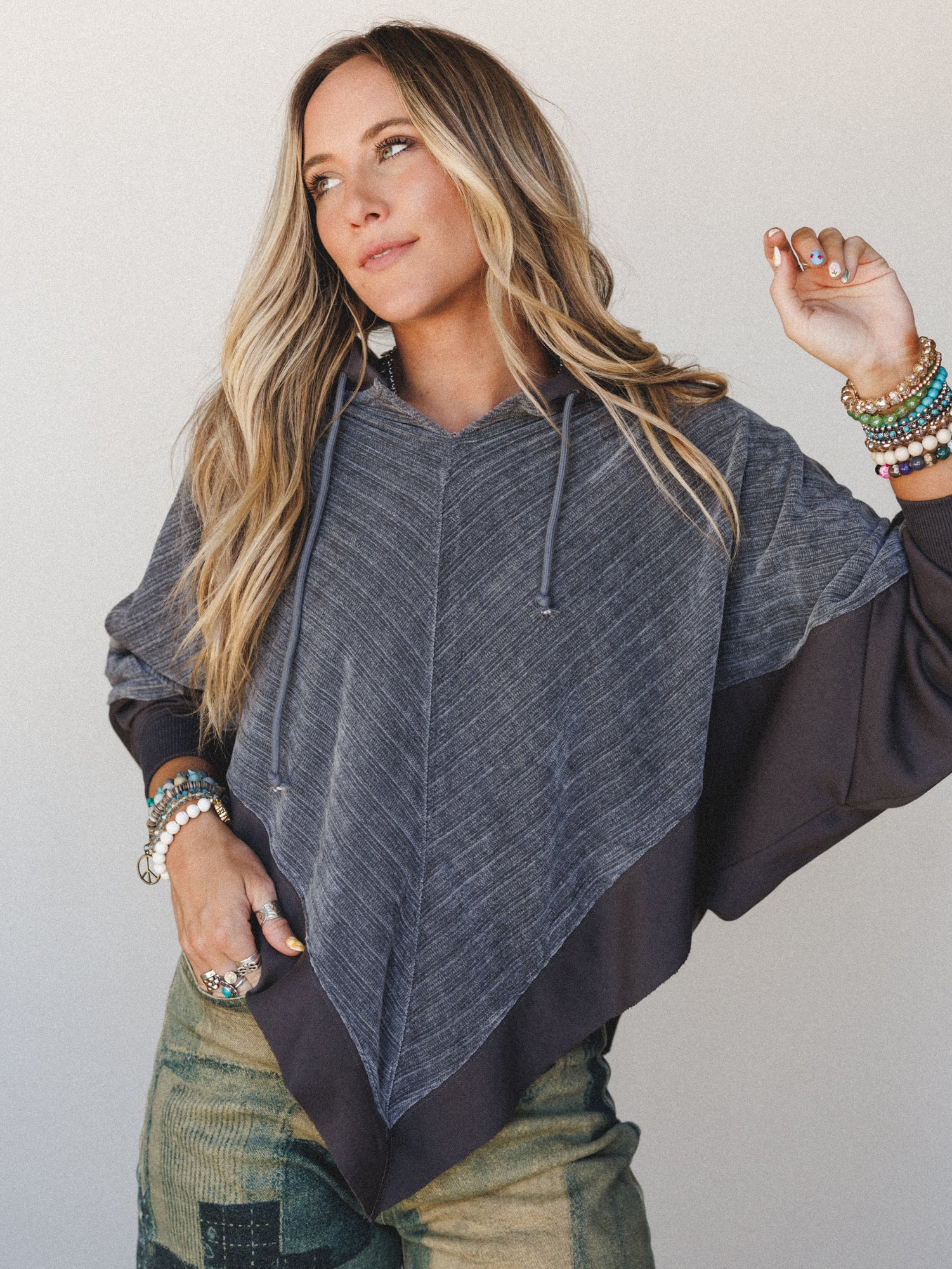 A relaxed, effortlessly styled model wears a charcoal color-blocked hoodie poncho with an asymmetric hem, layered over olive patchwork pants and stacked with beaded bracelets and silver rings — the kind of outfit that looks like you tried just enough