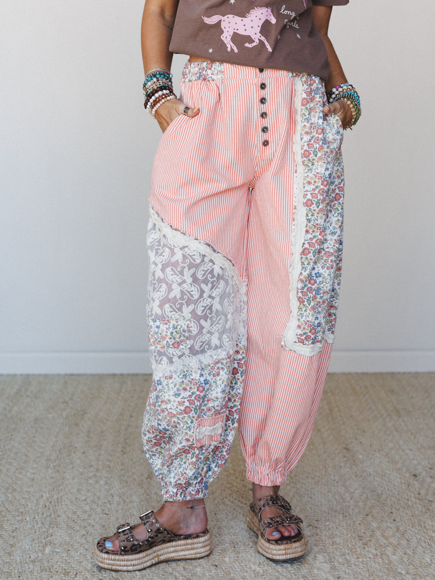 Patchwork harem-style pants in coral stripe mixed with floral and lace panels, styled with a graphic tee, stacked bead bracelets, and platform sandals — the kind of pants that have a whole personality before you even put them on.