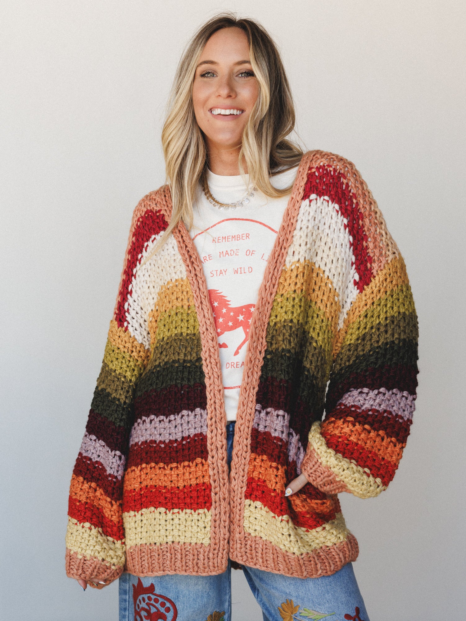 A chunky hand-knit striped cardigan in a full autumn rainbow of colors worn open over a graphic tee and embroidered jeans — the kind of piece that makes a whole outfit feel like a personality.