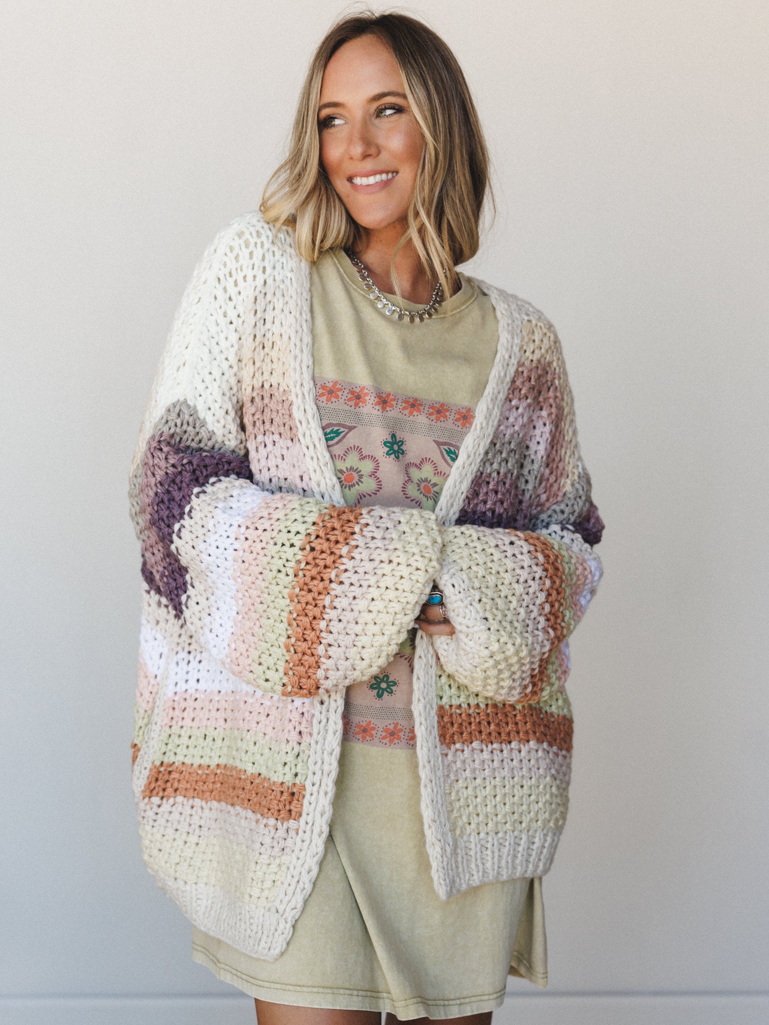 An oversized chunky knit cardigan in cream with multicolor stripes is layered over a sage green graphic tee and mini skirt — the kind of cozy, collected outfit that makes a Saturday feel intentional.