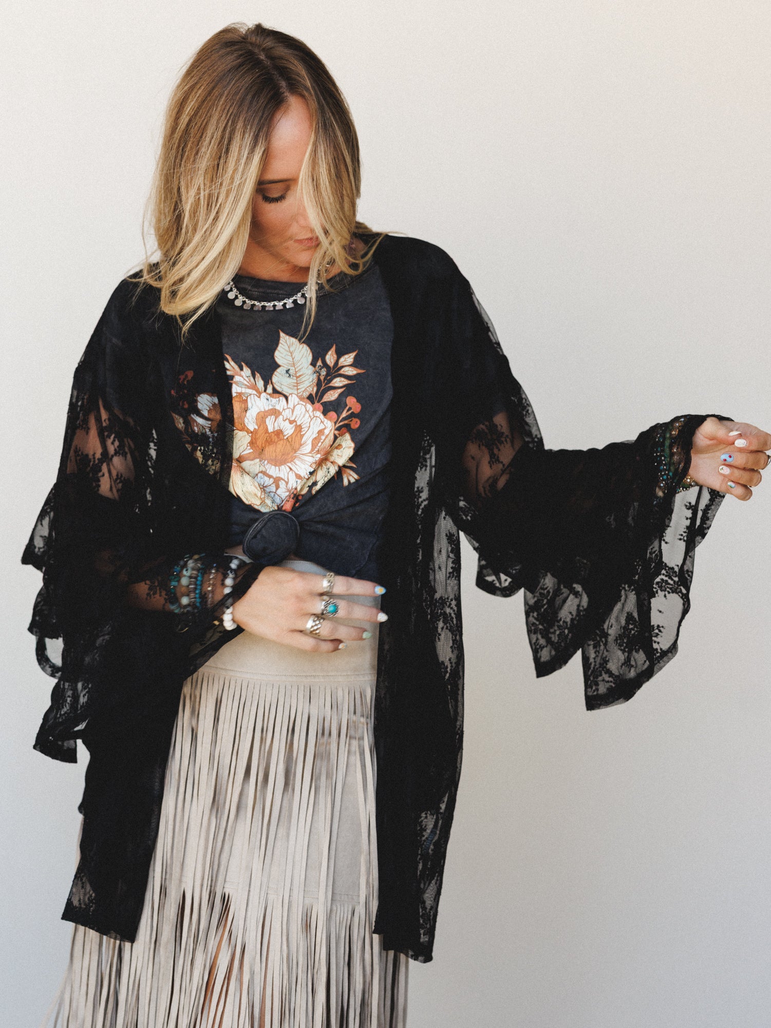 A layered boho outfit built around a black sheer lace kimono with dramatic bell sleeves, styled over a charcoal floral graphic tee and a cream fringe skirt — the kind of outfit that feels like it came together effortlessly but definitely didn't.