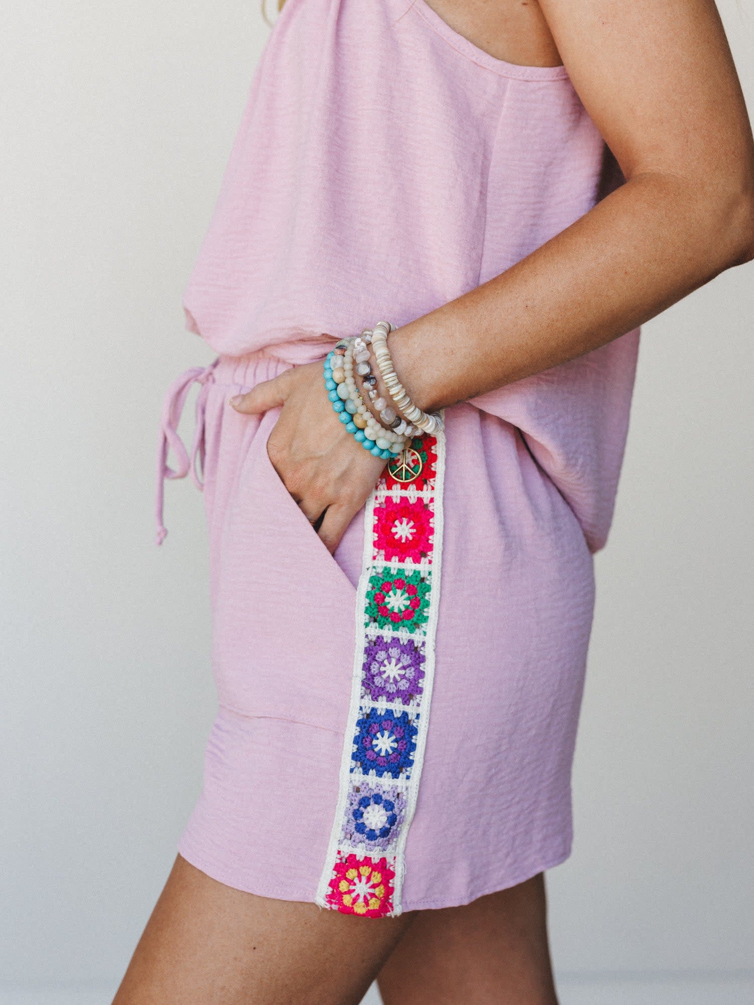 A close-up side shot of a lavender-pink romper or shorts set with a colorful granny square crochet trim running down the side seam, styled with a stack of beaded boho bracelets — the kind of outfit that feels like summer made it for you.