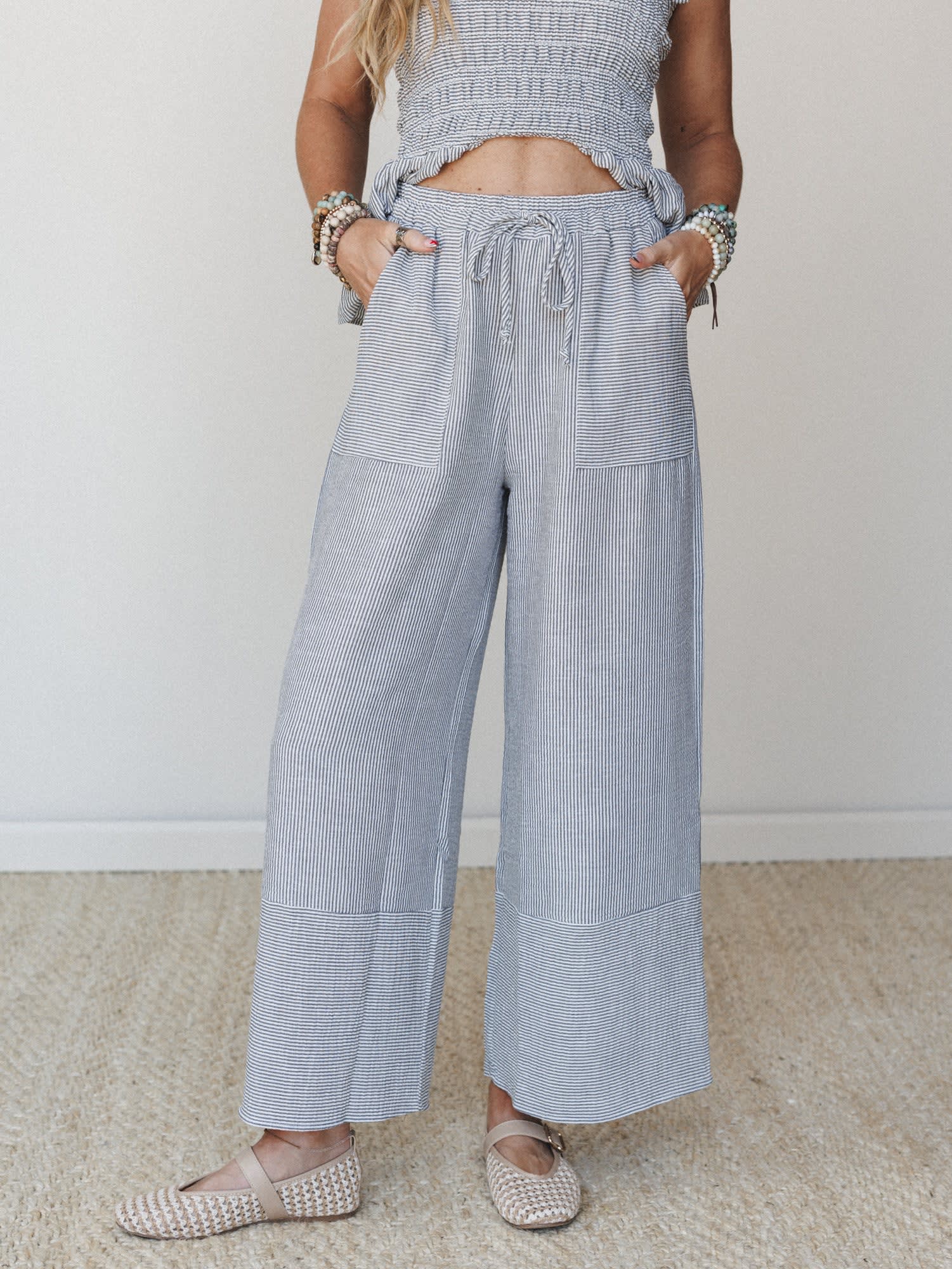 A cropped mid-shot of wide-leg striped pants with a smocked matching top peeking in at the waist — the kind of effortless set that looks pulled-together without trying too hard, styled with woven flats and stacked beaded bracelets.